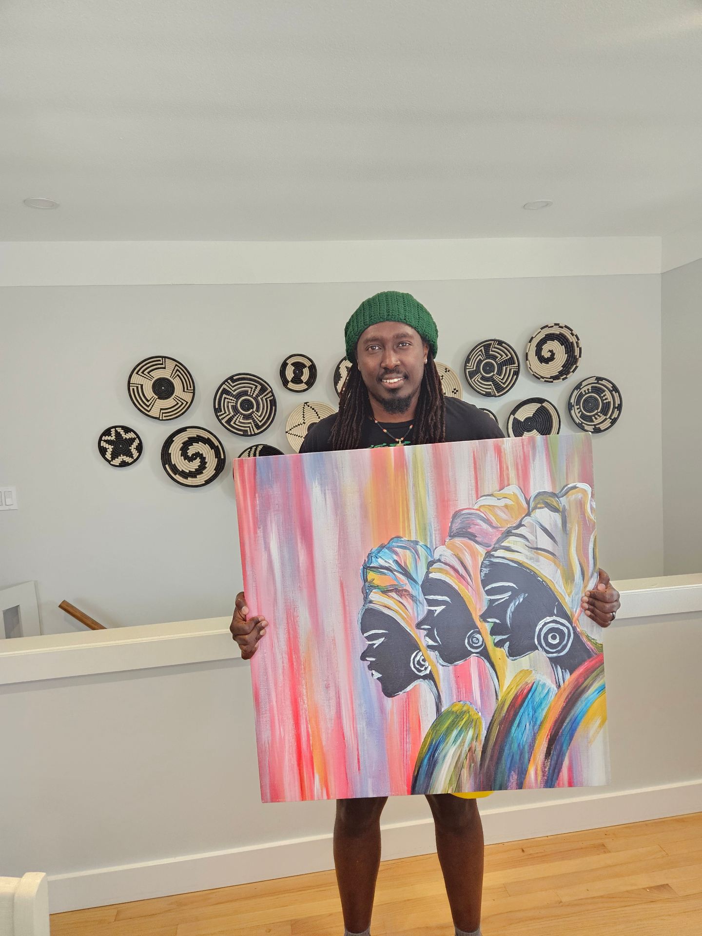 Celo holding a vibrant painting by the artist Ivan, featuring three silhouetted figures with colorful headwraps, in front of a wall of woven Rwandan baskets.